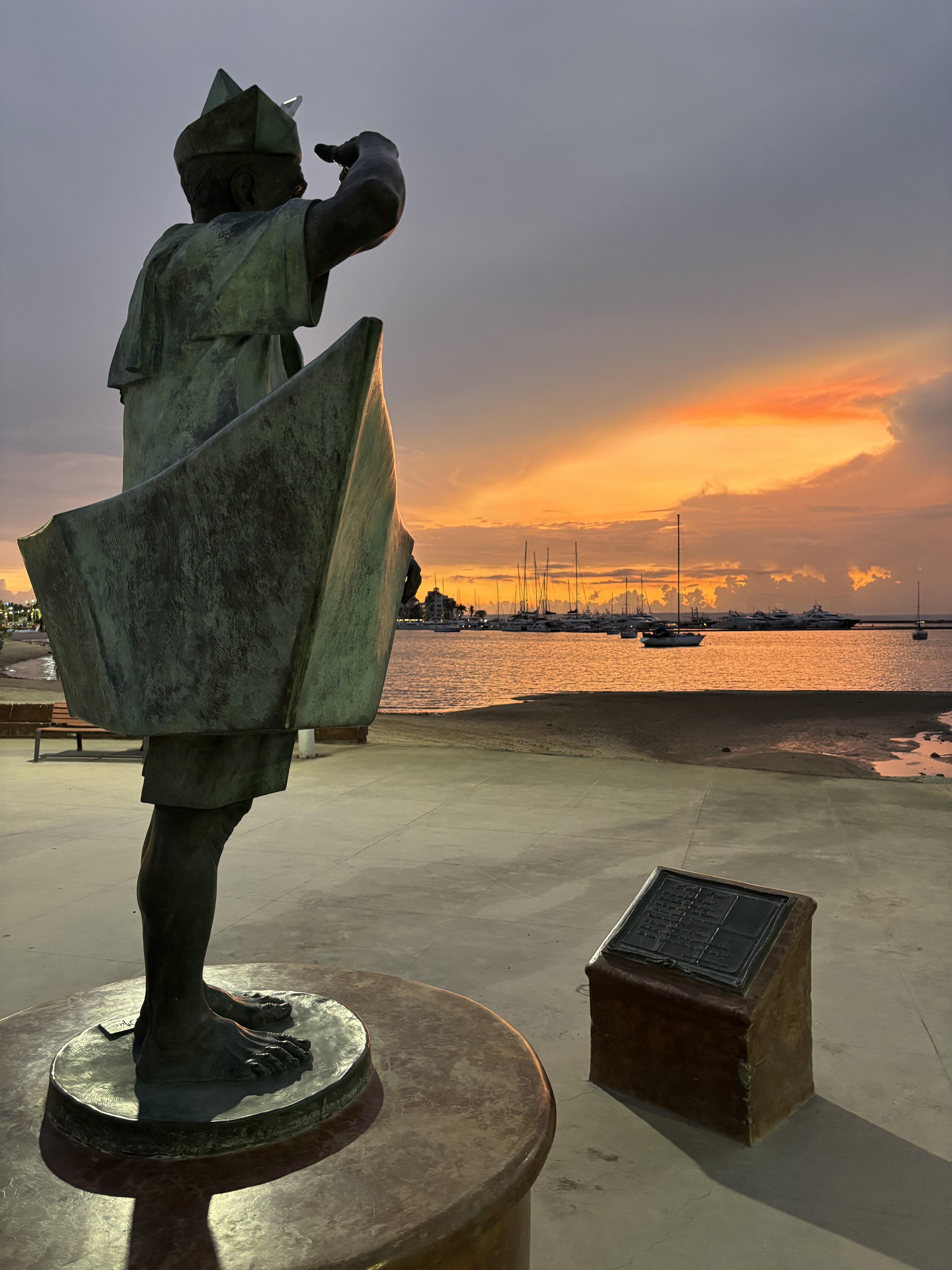 Aspiring Sailor Statue of a sailor overlooking sunset-lit marina and moored boats, capturing the spirit of adventure at sea.