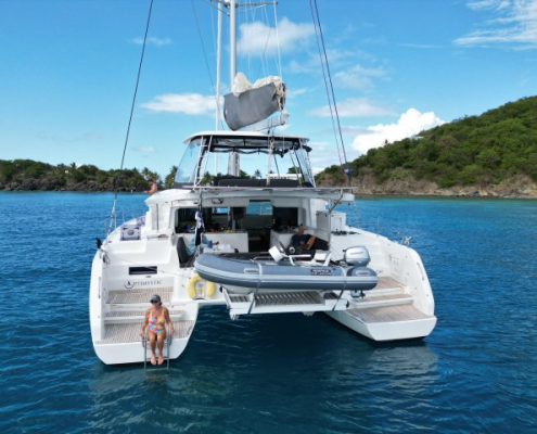 Catamaran anchored in turquoise waters near lush island with person relaxing on the swim ladder, enjoying a sunny day.