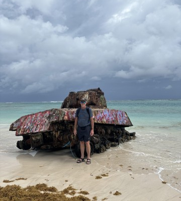 Man standing by a rusty, graffiti-covered tank stranded on a tropical beach with cloudy skies.